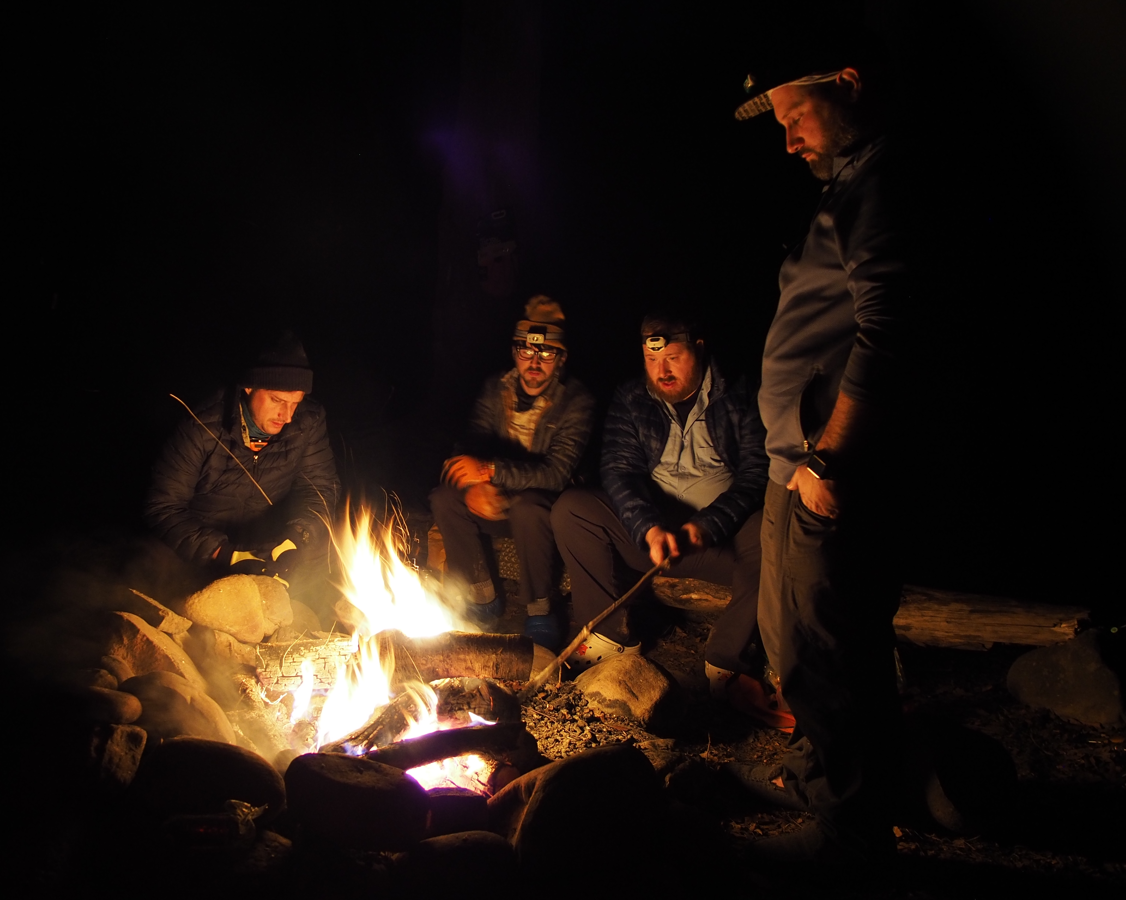 Four of the author's friends in cold weather clothing around a campfire at night. Three are seated, one is standing, all are staring into the fire.