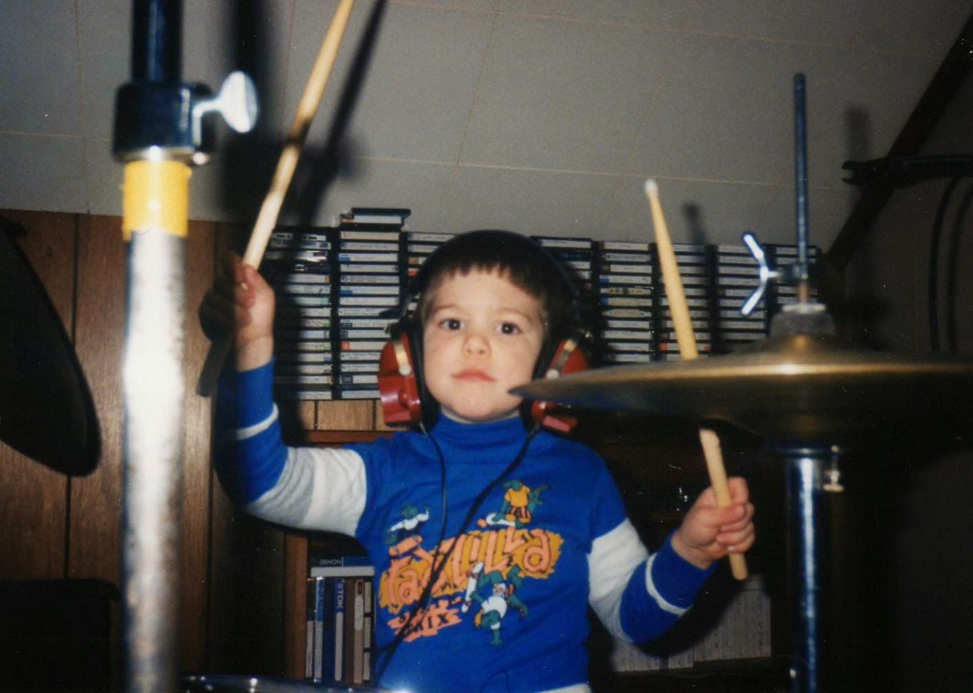 The author's younger brother, Chris, roughly 4 years old, sitting at a drum set, holding sticks, & wearing headphones.