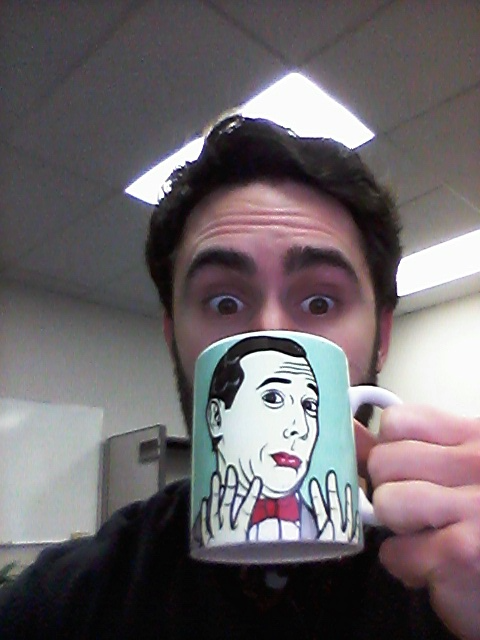 The author holding a coffee mug featuring the face & hands of PeeWee Herman, who is giving a gesture that seems to say 'Please don't talk to me'.