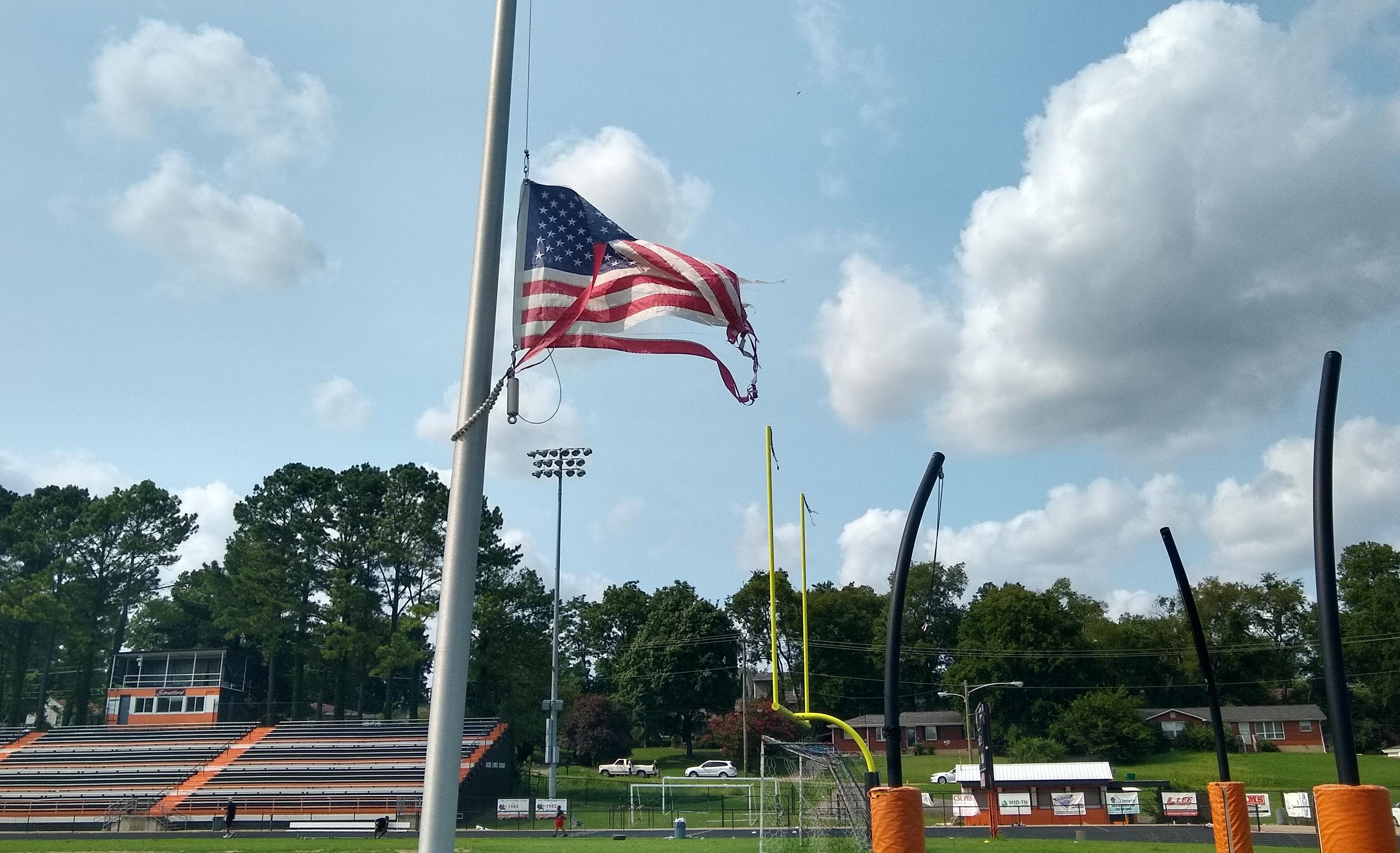 A slightly low-angle shot of a tattered American flag flying at half-staff in front of a high school football field.